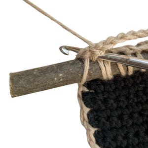 May include: Close-up of crochet work in progress. A silver crochet hook is inserted into a piece of wood, with beige yarn creating a chain stitch. The crochet project features black and beige yarn, showcasing the beginning of a textured pattern.