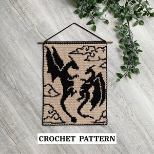 Puede incluir: Un tapiz de pared tejido a ganchillo con un diseño de dragones negros sobre un fondo beige. La obra de arte incluye dos dragones, nubes y el texto "CROCHET PATTERN" en la parte inferior. Se cuelga de una barra de madera con un cordón negro.