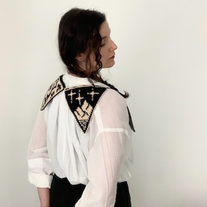 May include: A white button-down shirt with a black and beige crocheted collar. The collar features geometric patterns and cross designs. The shirt is paired with black bottoms.