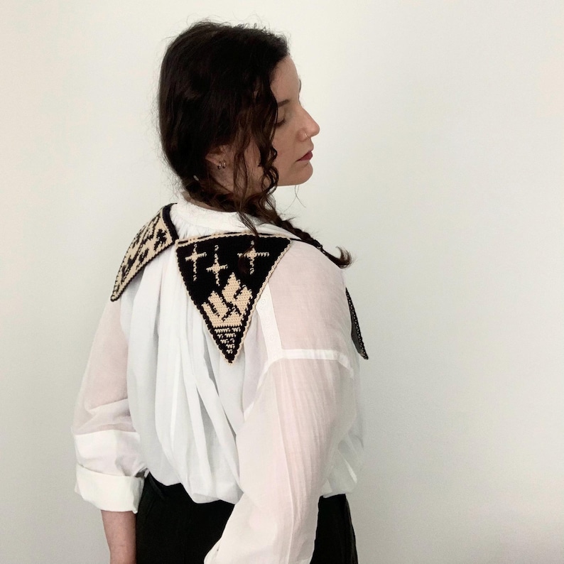 May include: A white button-down shirt with a black and beige crocheted collar. The collar features geometric patterns and cross designs. The shirt is paired with black bottoms.