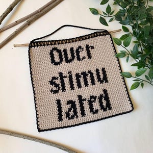 Digital crochet pattern for a wall tapestry featuring the word "Overstimulated" in bold black gothic-style lettering on a beige background. The pattern includes instructions to create the tapestry, which is bordered with black crochet edging. This listing is for the pattern only, not a finished item.