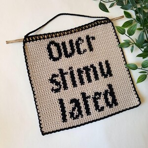 Digital crochet pattern for a wall tapestry featuring the word "Overstimulated" in bold black gothic-style lettering on a beige background. The pattern includes instructions to create the tapestry, which is bordered with black crochet edging. This listing is for the pattern only, not a finished item.