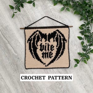 May include: A beige crochet wall hanging with a black heart-shaped design featuring bat wings and the words "bite me." The wall hanging is suspended from a wooden dowel and black cord. The text "CROCHET PATTERN" is at the bottom.