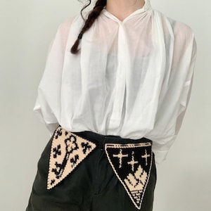 May include: Black shorts with two decorative, triangular crochet patches. One patch is black with a cream-colored design, and the other is cream-colored with a black design. The shorts are paired with a white, semi-sheer blouse.