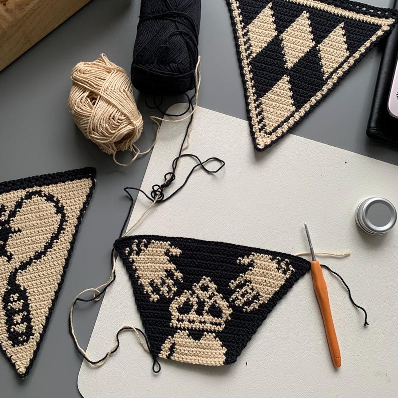 May include: A collection of crochet projects in progress, featuring black and beige yarn. Several triangular pieces with geometric and graphic designs are visible, along with balls of yarn and a crochet hook. The pieces are on a white surface.