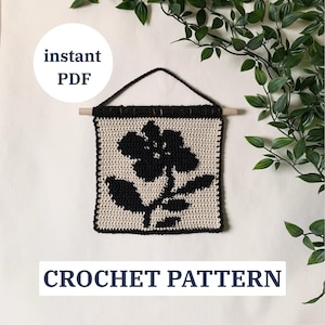 May include: A crocheted wall hanging featuring a black floral design on a cream background. The square piece is framed with black yarn and has a wooden dowel and black cord for hanging. The words "instant PDF" and "CROCHET PATTERN" are visible.