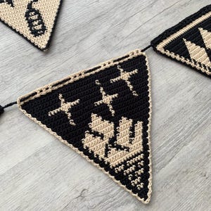 May include: A crocheted banner with triangular flags in black and cream. Each flag features a different design, including stars and geometric patterns. The banner is suitable for festive occasions or home decor.