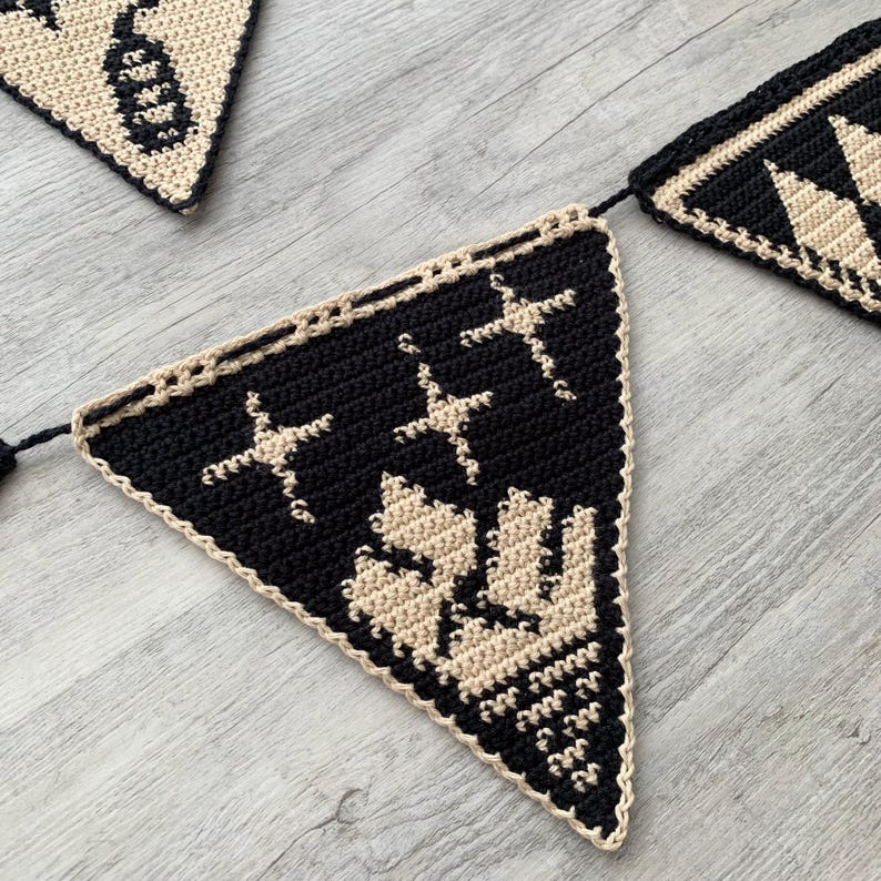 May include: A crocheted banner with triangular flags in black and cream. Each flag features a different design, including stars and geometric patterns. The banner is suitable for festive occasions or home decor.