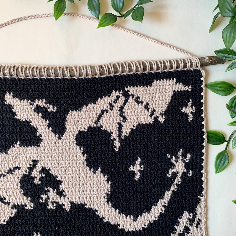 May include: A black and cream-colored crochet wall hanging with a map design. The piece is suspended from a natural wooden branch and a cream-colored cord. The map design features a detailed pattern, creating a striking contrast.