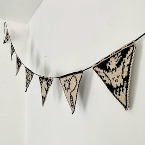May include: A black and beige crochet banner with triangular pennants. Each pennant features a unique black design on a beige background. The banner is strung on a black cord and hangs against a white wall.