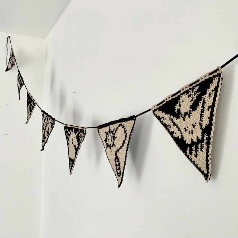 May include: A black and beige crochet banner with triangular pennants. Each pennant features a unique black design on a beige background. The banner is strung on a black cord and hangs against a white wall.