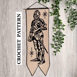 Op de afbeelding: Gehaakte wandhanger met een ridderontwerp in zwart en beige. De banner bevat de tekst "CROCHET PATTERN" aan de linkerkant. De ridder wordt afgebeeld met een zwaard en sterren. Het stuk is ontworpen om op te hangen.