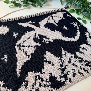 May include: A black and beige crochet wall hanging featuring a dragon design. The dragon is depicted in beige against a black background. The wall hanging is attached to a wooden dowel with a beige cord for hanging.