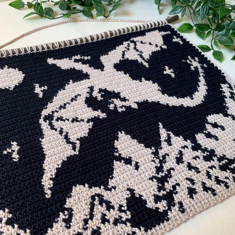 May include: A black and beige crochet wall hanging featuring a dragon design. The dragon is depicted in beige against a black background. The wall hanging is attached to a wooden dowel with a beige cord for hanging.