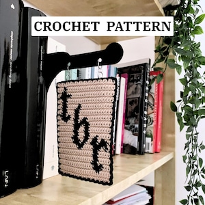 Könnte beinhalten: Beige gehäkelter Lesezeichen mit schwarzen Buchstaben "tbr" und schwarzem Rand, an einem schwarzen Metallhaken aufgehängt. Der Text "CROCHET PATTERN" ist im Bild zu sehen. Bücher stehen auf einem Holzregal.