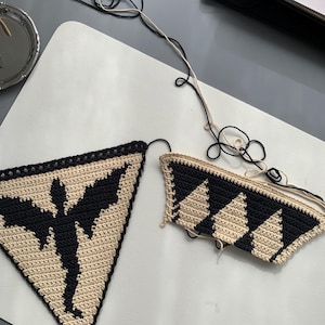 May include: Two handmade crochet pieces. One is a triangle with a black bat silhouette on a tan background. The other is a partial triangle with black and tan diamond pattern. Black and tan yarn is attached.