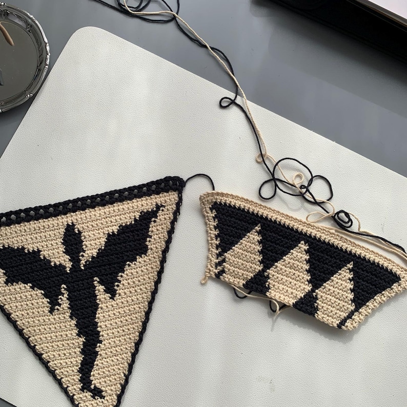 May include: Two handmade crochet pieces. One is a triangle with a black bat silhouette on a tan background. The other is a partial triangle with black and tan diamond pattern. Black and tan yarn is attached.