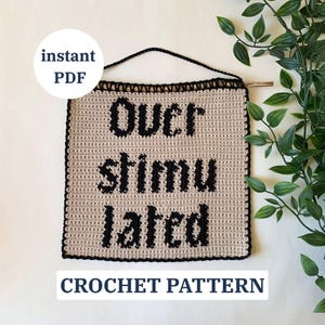 May include: A beige crochet wall hanging with the words "Over Stimulated" in black block letters. The sign is framed with a black border and has a black hanging cord. The words "instant PDF" and "CROCHET PATTERN" are also visible.