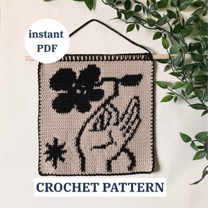 May include: A crocheted wall hanging featuring a black flower and hand design on a beige background. The piece is square, with a black border and a wooden dowel for hanging. Text on the image reads "instant PDF" and "CROCHET PATTERN".
