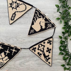 May include: A handmade crochet banner with triangular flags. Each flag features a black design on a tan background. The designs include a key, a unicorn, and other symbols. The banner is strung on a black cord.