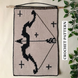 Może przedstawiać: Beżowy szydełkowy gobelin ścienny z czarnym wzorem łuku i strzały oraz czterema czarnymi krzyżami. Tekst "CROCHET PATTERN" jest wydrukowany pionowo po prawej stronie. Zawieszony na drewnianym drążku z czarnym sznurkiem.