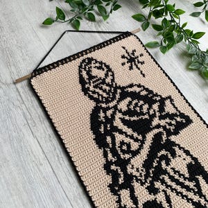 May include: A beige and black crochet wall hanging featuring a stylized figure and starburst design. The rectangular piece is suspended from a wooden dowel with a black cord. The edges are finished with a black border.