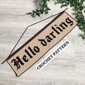 May include: A beige crochet wall hanging with the words "Hello Darling" in black gothic lettering. The banner is suspended from a wooden dowel with a black string. The words "CROCHET PATTERN" are also visible.
