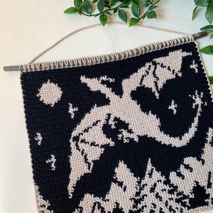 May include: A handmade crochet wall hanging featuring a dragon design. The dragon is light beige against a dark navy blue background. The wall hanging is attached to a wooden dowel with a beige cord for hanging. The design includes a moon and stars.