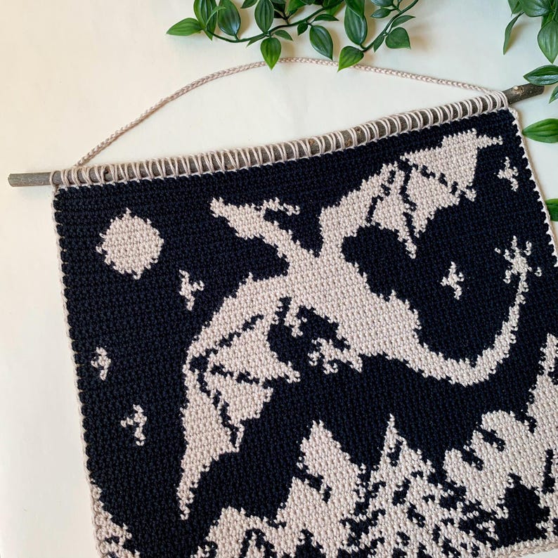 May include: A handmade crochet wall hanging featuring a dragon design. The dragon is light beige against a dark navy blue background. The wall hanging is attached to a wooden dowel with a beige cord for hanging. The design includes a moon and stars.