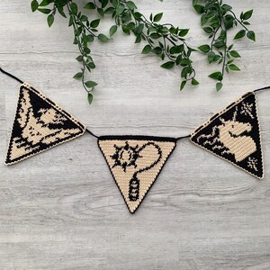 May include: A crocheted banner with three triangular flags. Each flag features a black design on a tan background. The designs include a creature, a sun-like symbol with a question mark, and a unicorn. The banner is strung on a black cord.