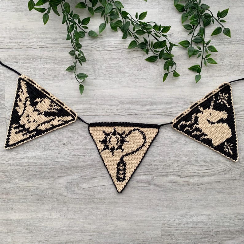 May include: A crocheted banner with three triangular flags. Each flag features a black design on a tan background. The designs include a creature, a sun-like symbol with a question mark, and a unicorn. The banner is strung on a black cord.