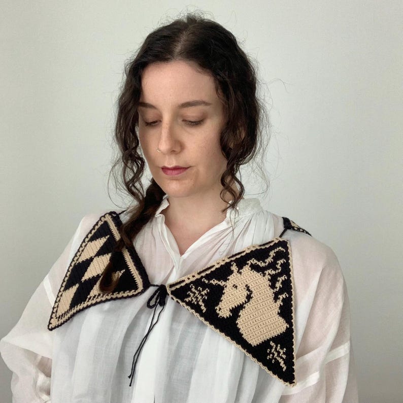 May include: A black and cream-colored crochet collar with a geometric and unicorn design. The collar is triangular and tied with a black string. The collar is worn over a white blouse.