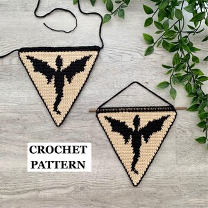 Pode incluir: Dois tapeçarias triangulares de crochê com uma silhueta de dragão preto em um fundo bege. Um estandarte pende de um cordão de lã preta e o outro está suspenso em uma vareta de madeira com um cordão preto. O texto diz CROCHET PATTERN.