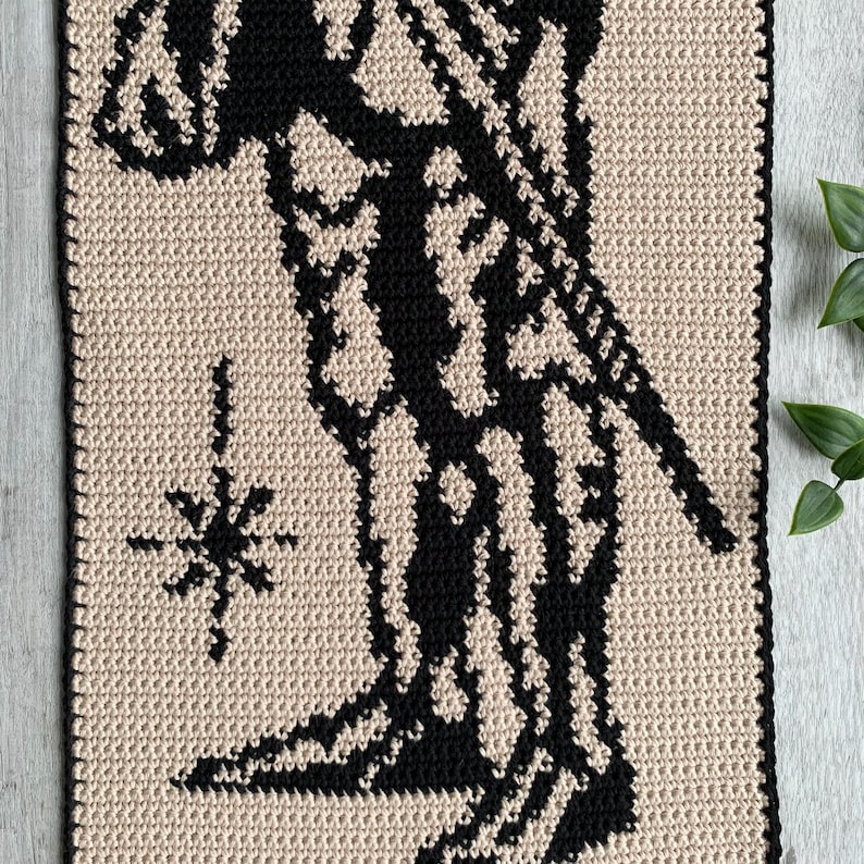 May include: A crochet wall hanging featuring a black silhouette design on a beige background. The design includes a figure with a star-like shape. The piece is handmade and has a textured appearance.