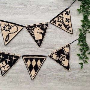 May include: A black and beige crochet banner with triangular flags. Each flag features a different design, including a unicorn, a sun, and geometric patterns. The banner is strung on a black cord and is suitable for home decor.