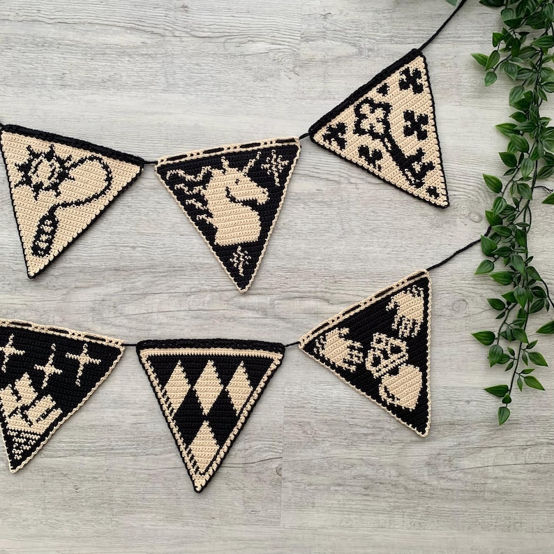 May include: A black and beige crochet banner with triangular flags. Each flag features a different design, including a unicorn, a sun, and geometric patterns. The banner is strung on a black cord and is suitable for home decor.