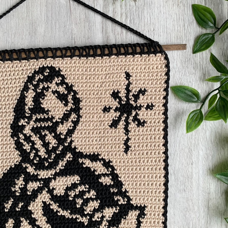 May include: A square, beige crochet wall hanging with a black border and a black design. The design features a figure with a hood and a snowflake. The wall hanging is suspended by a black cord and a wooden dowel.