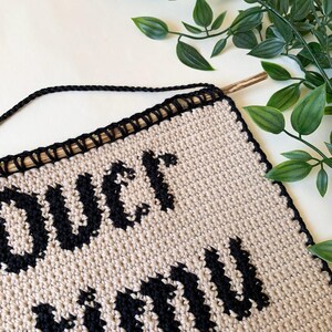 Digital crochet pattern for a wall tapestry featuring the word "Overstimulated" in bold black gothic-style lettering on a beige background. The pattern includes instructions to create the tapestry, which is bordered with black crochet edging. This listing is for the pattern only, not a finished item.