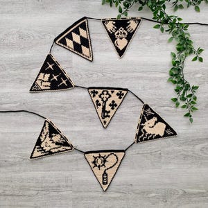 May include: A black and cream-colored triangle banner with various symbols, including a key, a star, and a teapot. The banner is strung on a black cord and is displayed against a light gray wooden background.