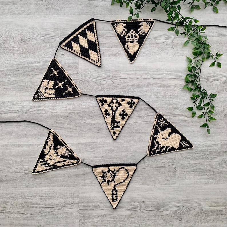May include: A black and cream-colored triangle banner with various symbols, including a key, a star, and a teapot. The banner is strung on a black cord and is displayed against a light gray wooden background.