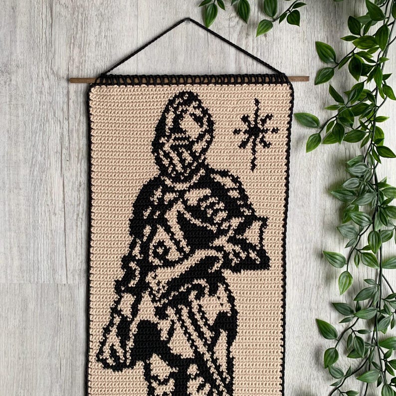 May include: A crochet wall hanging featuring a black and beige design of a knight in armor. The knight is holding a sword. A star is to the right of the knight. The wall hanging is rectangular and has a black border.