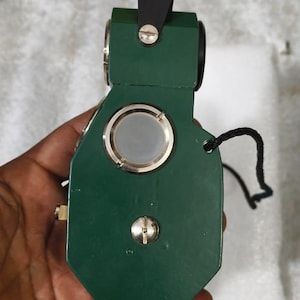 May include: A dark green handheld geological compass featuring a circular glass lens, a metallic adjustment screw, and a black strap. The device has a sturdy, angular frame with a top-mounted black tab and a silver screw fastener on the front.