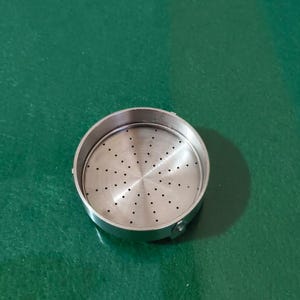 Może przedstawiać: Okrągłe narzędzie kuchenne ze stali nierdzewnej z perforowanym wnętrzem. Ma podniesioną, zaokrągloną krawędź i jest umieszczone na zielonej powierzchni. Wnętrze ma małe, równomiernie rozmieszczone otwory.