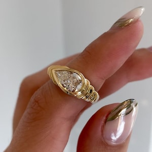 May include: A gold ring with a pear-shaped diamond center stone. The ring has a textured band with a ribbed design. The diamond is set in a bezel setting. The ring is shown on a hand with manicured nails.