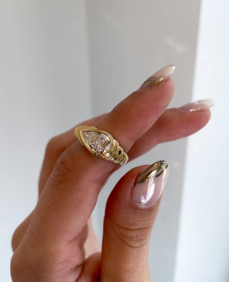 May include: A gold ring with a pear-shaped diamond center stone. The ring has a textured band with a ribbed design. The diamond is set in a bezel setting. The ring is shown on a hand with manicured nails.