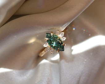 May include: A gold ring featuring a large marquise-cut dark green gemstone as the centerpiece, flanked by a cluster of smaller, clear, pear-shaped stones on both sides. The band is smooth and polished, reflecting light with a bright, metallic sheen.