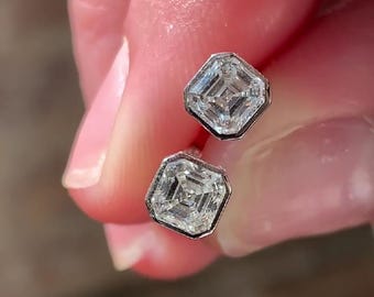 May include: A pair of stud earrings featuring square-shaped, step-cut clear gemstones set in polished silver-toned metal bezels. Each stone displays a concentric, geometric facet pattern that reflects light, creating a bright, multi-layered appearance.