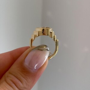 May include: A gold-colored ring with a geometric design. The ring features a series of stepped elements around the band and a central, possibly gemstone, setting. The ring is held up, showcasing its design and craftsmanship.