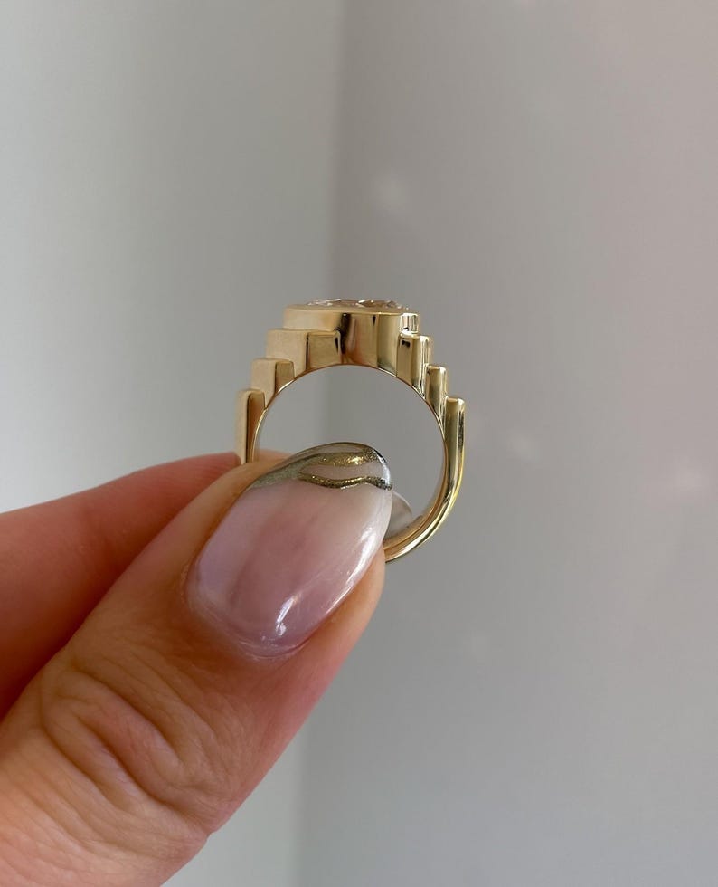 May include: A gold-colored ring with a geometric design. The ring features a series of stepped elements around the band and a central, possibly gemstone, setting. The ring is held up, showcasing its design and craftsmanship.