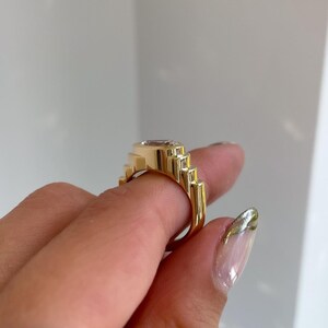 May include: A gold-colored ring with a rectangular, light-colored gemstone. The ring has a stepped design on the sides, creating a geometric pattern. The ring is held up by a person's fingers.
